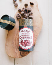 Load image into Gallery viewer, Opened tube of Chocolate Coated Cherries with the chocolate coated cherries spilling out of the top on a wood cutting board.