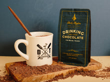 Load image into Gallery viewer, Belize Drinking Chocolate bag next to a Dick Taylor Chocolate branded diner much with a stirrer in it set on a wood cutting board with drinking chocolate mix sprinkled around.