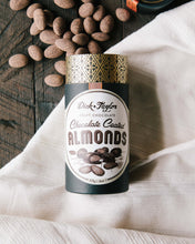 Load image into Gallery viewer, Opened tube of Chocolate Coated Almonds with the chocolate coated almonds spilling out of the top of the tube on a wood background with a white cloth.