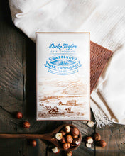 Load image into Gallery viewer, Hazelnut bar packing on top of an unpackaged chocolate bar on a wooden background decorated with a white cloth and hazelnuts.