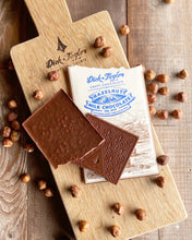 Load image into Gallery viewer, Hazelnut bar broken in half on top of hazelnut bar packaging on a wooden cutting board with hazelnuts scattered around.