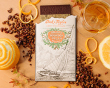 Load image into Gallery viewer, An opened Orange Bourbon Pecan bar on a wooden background decorated with chopping pecans, orange peels, and a glass of bourbon.