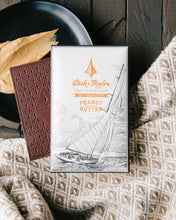 Load image into Gallery viewer, Peanut Butter bar packaging on top of an unpackaged chocolate bar on a wooden background. Decorated with a tan and white cloth and a plate with a smear of peanut butter.