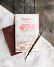 Load image into Gallery viewer, Vanilla Milk bar packaging with a vanilla bean on top, on top of a unpackaged chocolate bar on a wooden background with a white cloth.