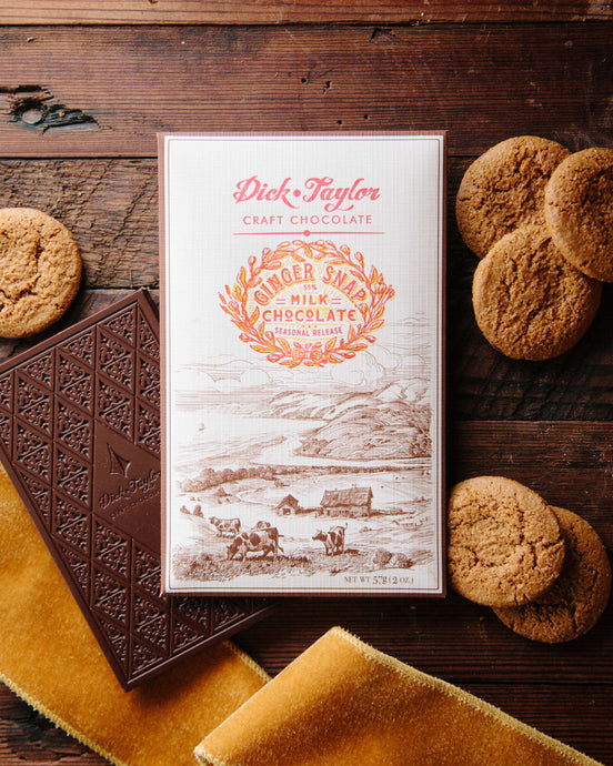 Ginger Snap bar packaging on top of an unpackaged chocolate bar on a wooden background decorated with ginger snap cookies and an orange ribbon.