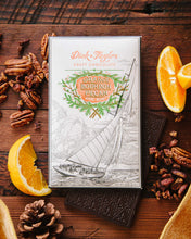 Load image into Gallery viewer, Orange Bourbon Pecan bar packaging on top of a unpackaged chocolate bar on a wooden background decorated with orange slices and chopped pecans.
